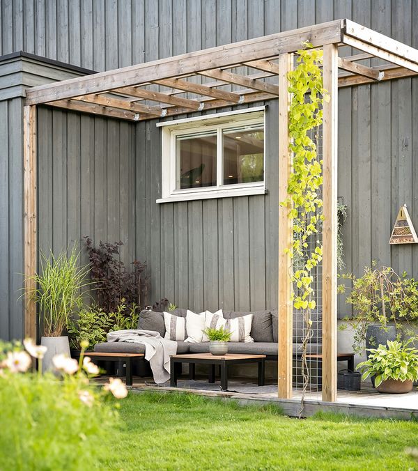Pergola aus Holz mit Sofa-Sitzecke vor gräulich gestrichener Holzfassade eines Hauses.