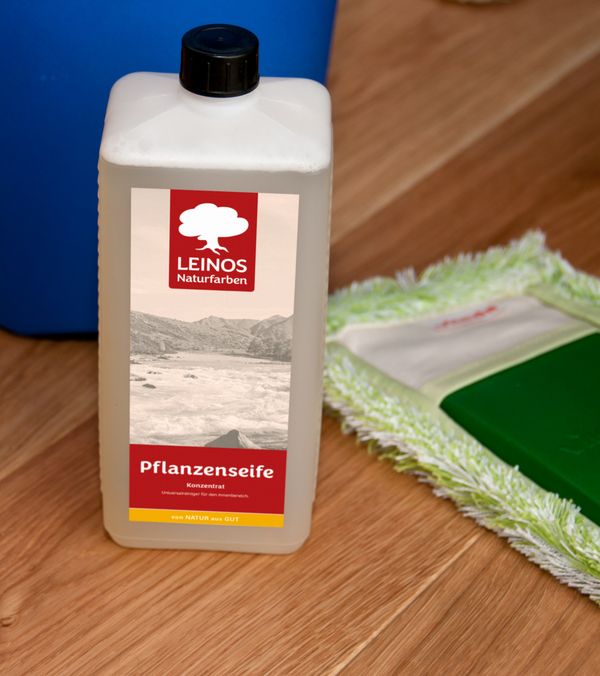 1l Gebinde der LEINOS Pflanzenseife, blauer Eimer und Wischmopp auf Parkett.
