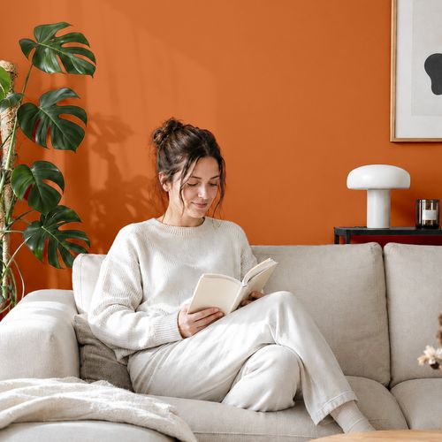 Frau liest ein Buch auf einem Sofa vor einer mit der Farbe "Abendrot" gestrichenen Wand