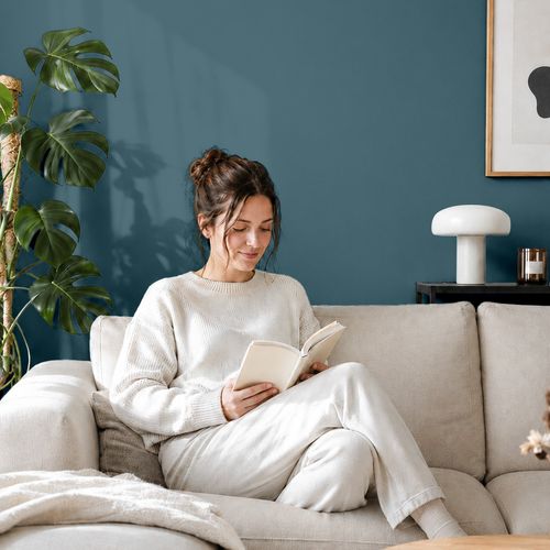 Frau liest ein Buch auf einem Sofa vor einer mit der Farbe "Atlantikblau" gestrichenen Wand