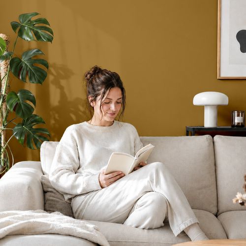Frau liest ein Buch auf einem Sofa vor einer mit der Farbe "Bernsteingelb" gestrichenen Wand