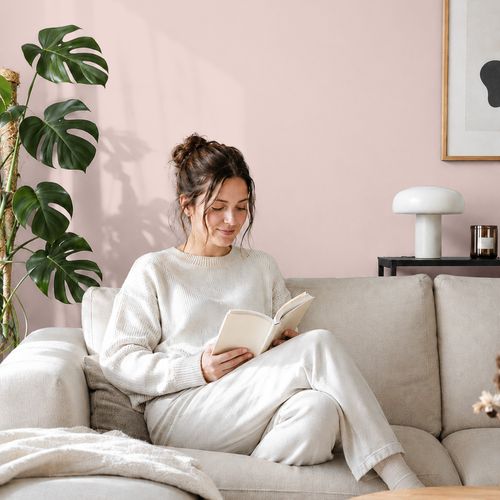 Frau liest ein Buch auf einem Sofa vor einer mit der Farbe "Blütenrosa" gestrichenen Wand