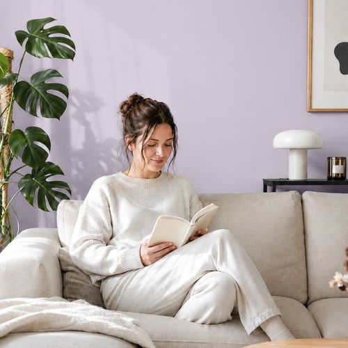 Frau liest ein Buch auf einem Sofa vor einer mit der Farbe "Fliederduft" gestrichenen Wand