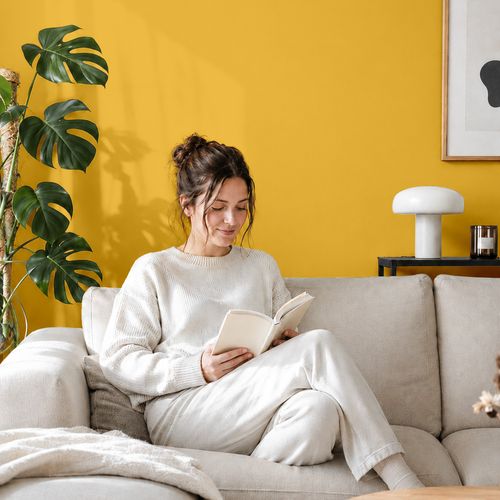 Frau liest ein Buch auf einem Sofa vor einer mit der Farbe "Goldene Stunde" gestrichenen Wand