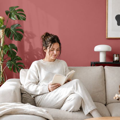 Frau liest ein Buch auf einem Sofa vor einer mit der Farbe "Hibiskus Rot" gestrichenen Wand
