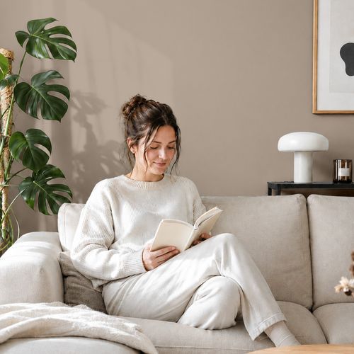 Frau liest ein Buch auf einem Sofa vor einer mit der Farbe "Lehmsand" gestrichenen Wand