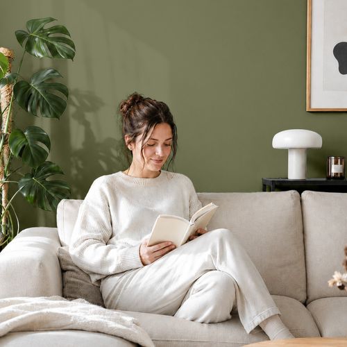 Frau liest ein Buch auf einem Sofa vor einer mit der Farbe "Moosgrün" gestrichenen Wand