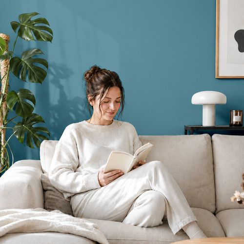 Frau liest ein Buch auf einem Sofa vor einer mit der Farbe "Ozeanblau" gestrichenen Wand