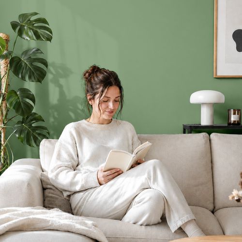 Frau liest ein Buch auf einem Sofa vor einer mit der Farbe "Salbeigrün" gestrichenen Wand