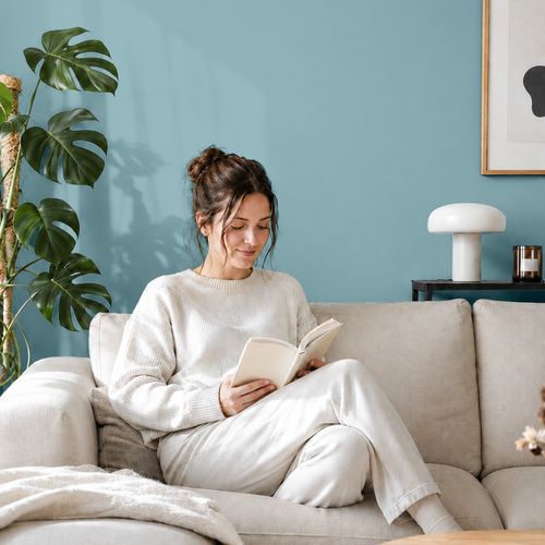 Frau liest ein Buch auf einem Sofa vor einer mit der Farbe "Sommerhimmel" gestrichenen Wand